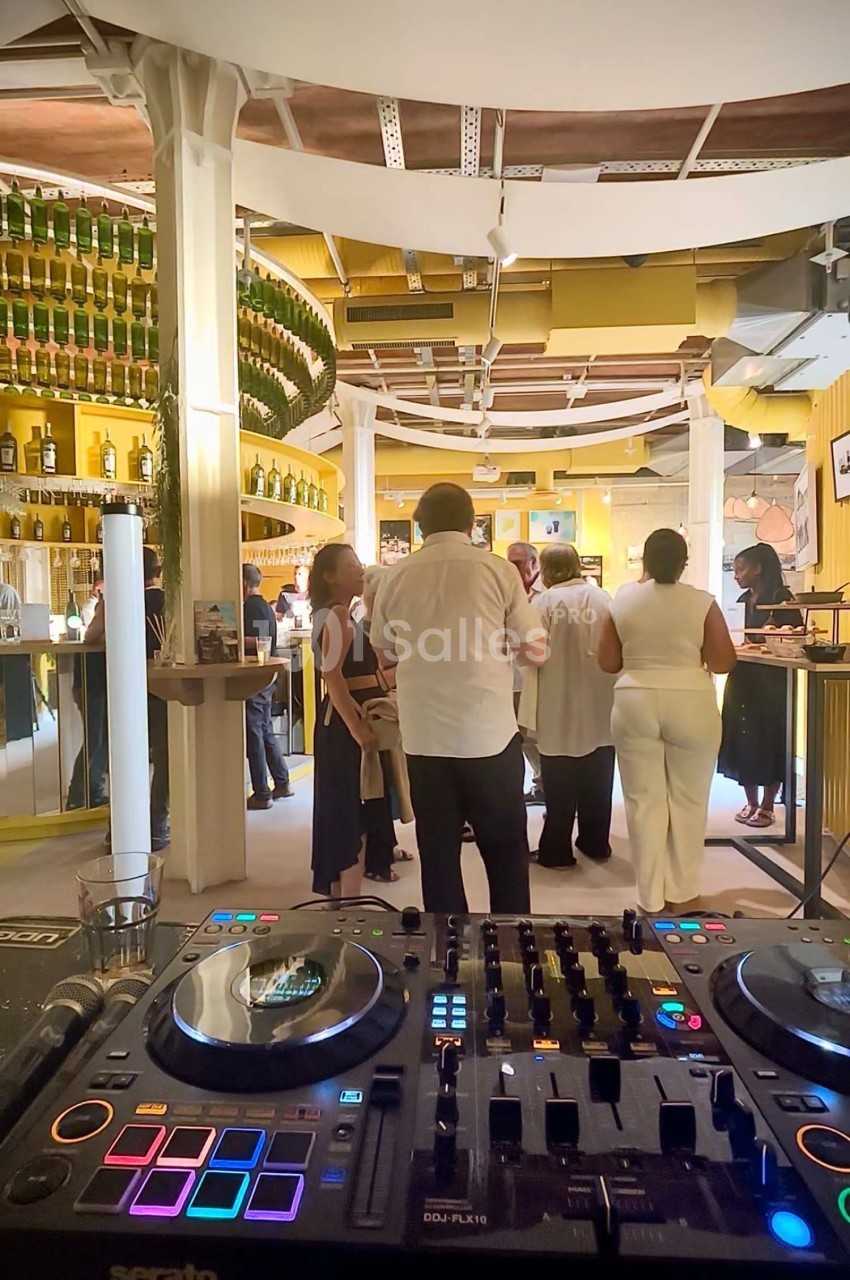 Vue d'une salle animée avec des personnes discutant près d'un bar, en arrière-plan d'une table de mixage DJ.