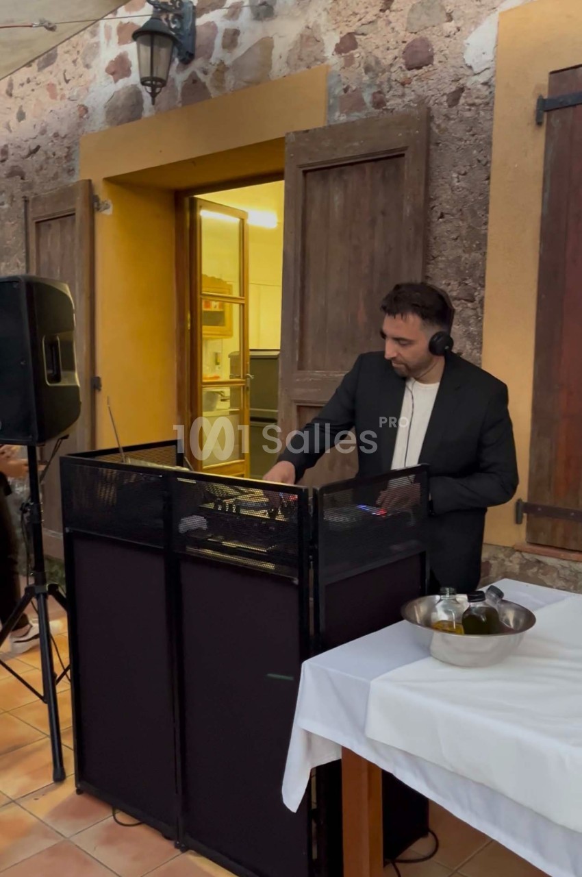 Un DJ en costume mixe de la musique sur une console dans une cour avec des murs en pierre et des tables dressées.