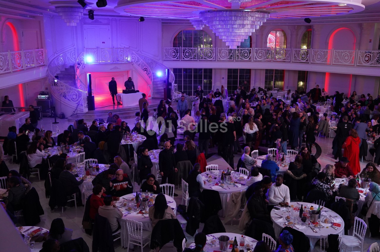Salle de réception éclairée en violet et rouge, remplie d'invités assis à des tables rondes lors d'un événement.