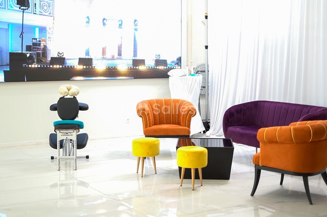 Salon moderne avec fauteuils colorés, tabourets jaunes, table basse noire et écran lumineux en arrière-plan.