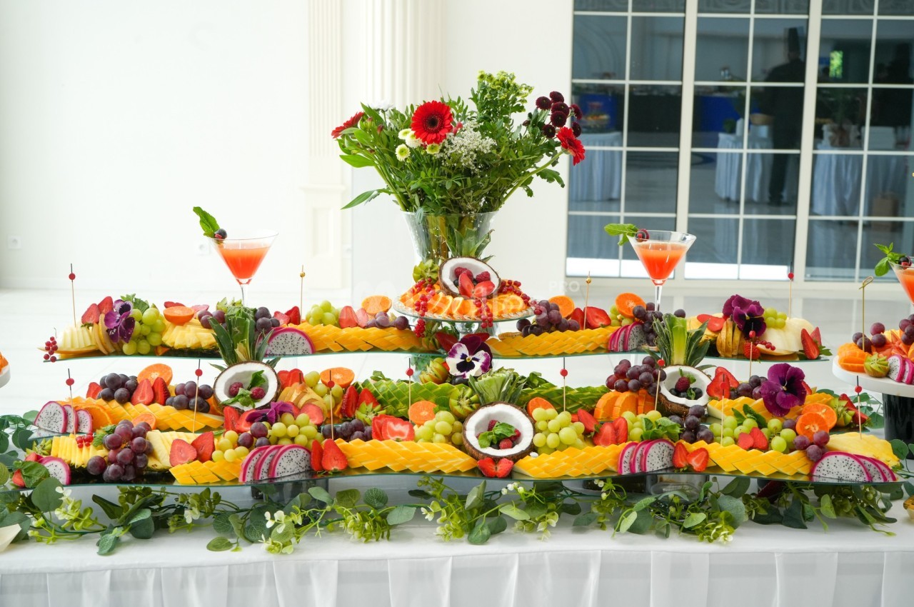Buffet décoratif avec des fruits variés, disposés en étages, entouré de fleurs et de cocktails colorés.