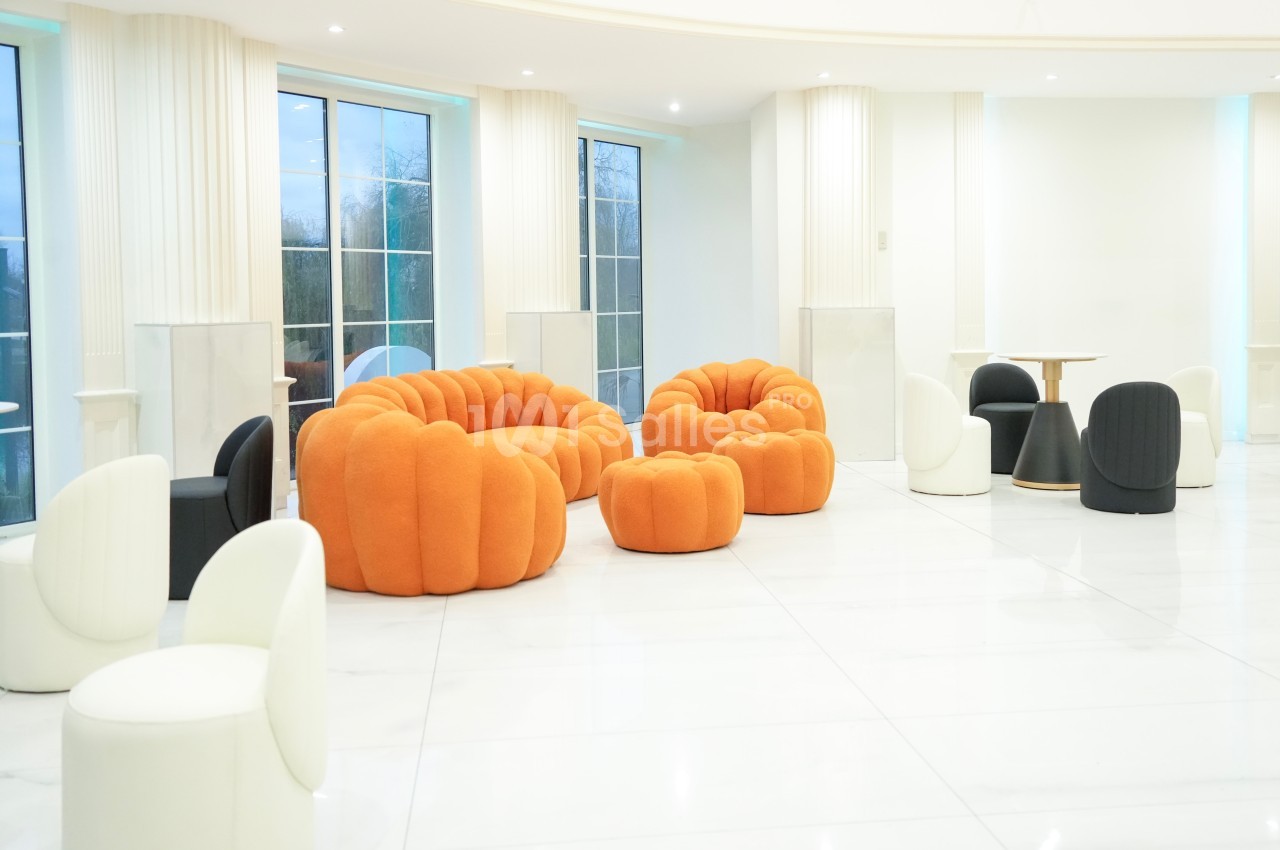 Salle lumineuse avec mobilier design comprenant des fauteuils orange et noirs, entourés de grandes fenêtres.
