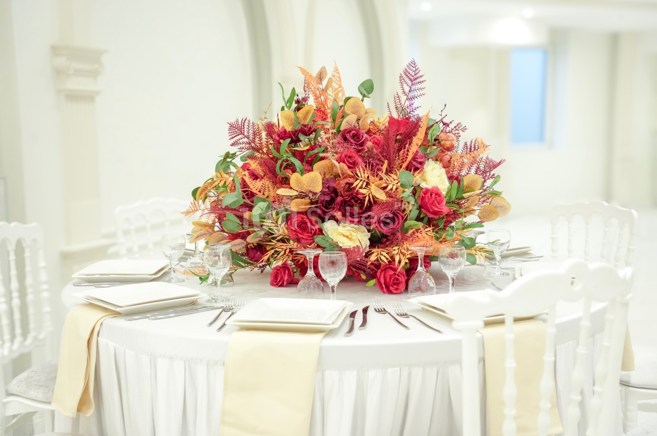 Centre de table floral aux tons rouges, jaunes et orangés, disposé sur une table dressée avec nappes blanches et serviettes…