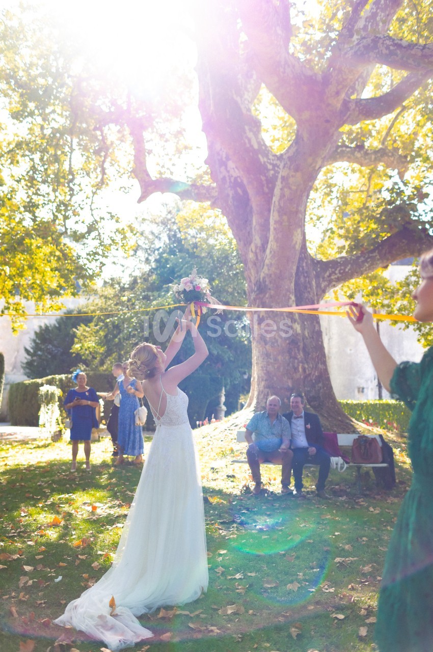Une mariée lance son bouquet dans un jardin ensoleillé, entourée d'invités sous un grand arbre.