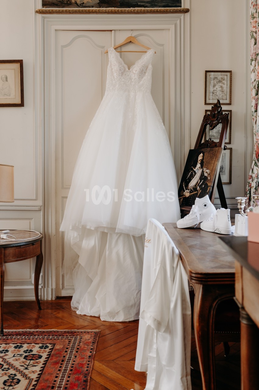 Robe de mariée blanche suspendue à une porte dans une pièce élégante avec parquet et meubles anciens.