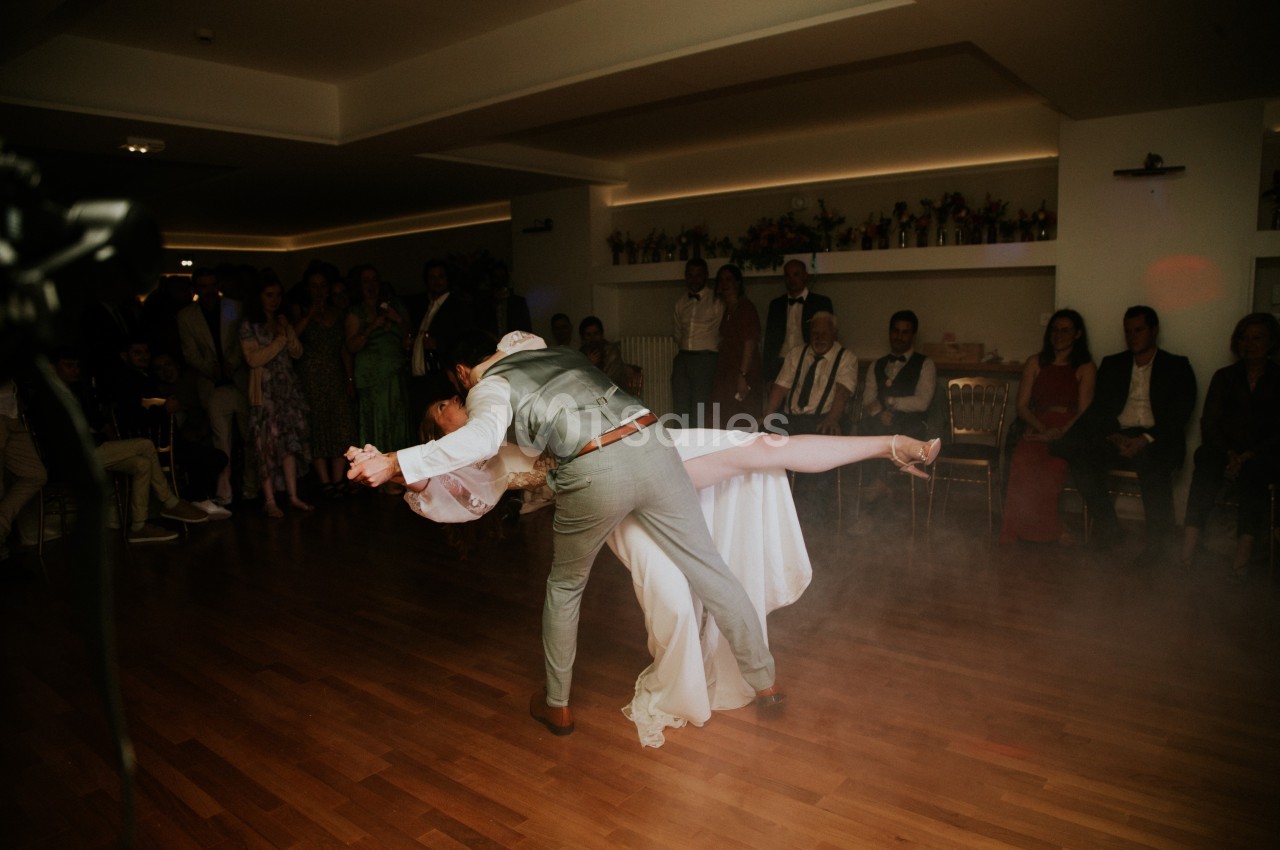 Un couple effectue une danse de mariage avec une figure élégante sur une piste en bois, entouré d'invités attentifs.