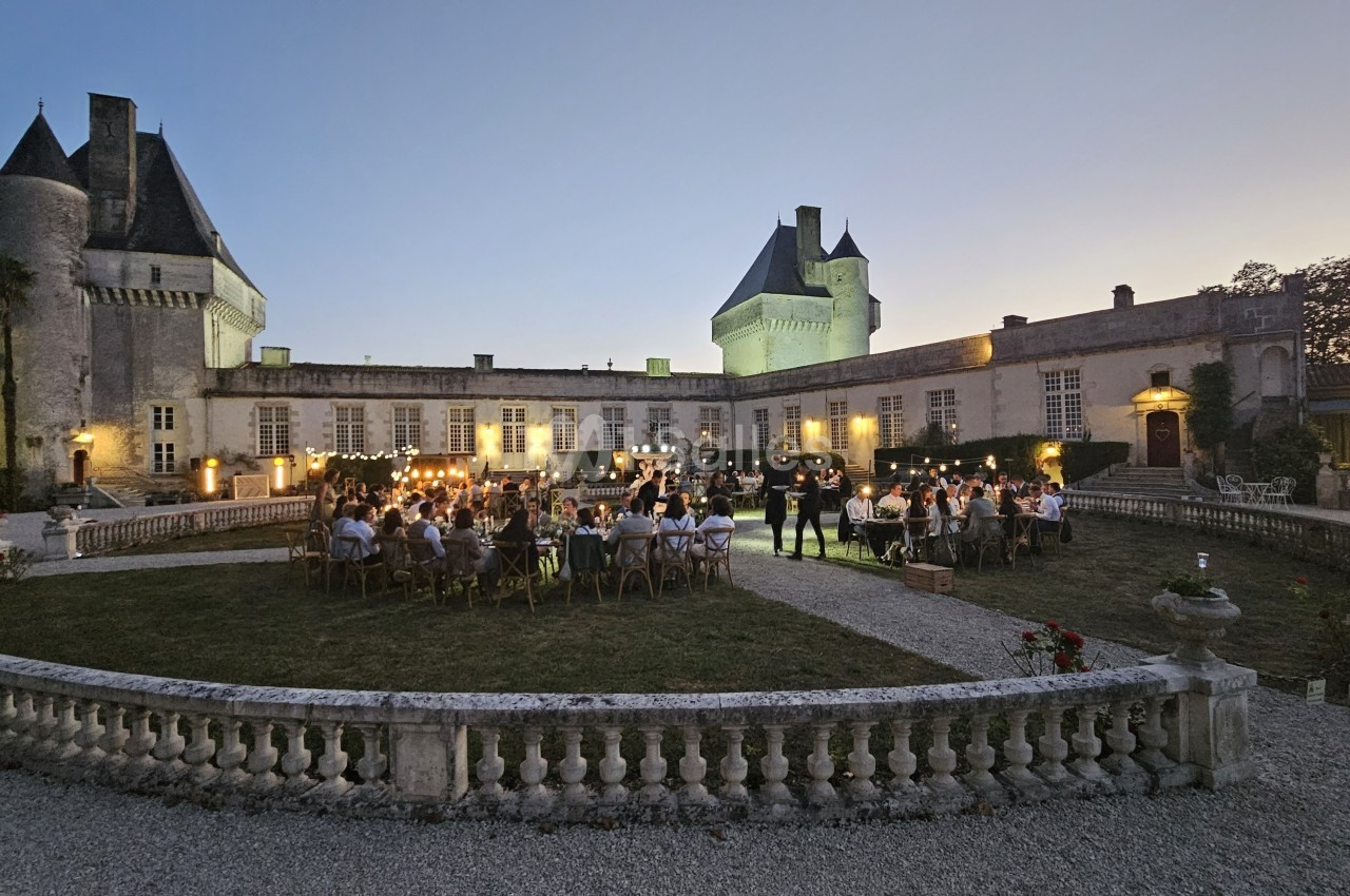 Dîner en plein air au crépuscule dans la cour d'un château illuminé, entouré de jardins et d'invités.