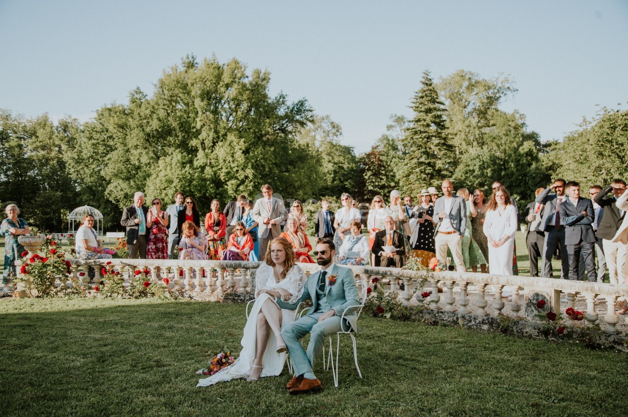 Un couple assis lors d'une cérémonie en plein air, entouré d'invités debout dans un jardin fleuri.