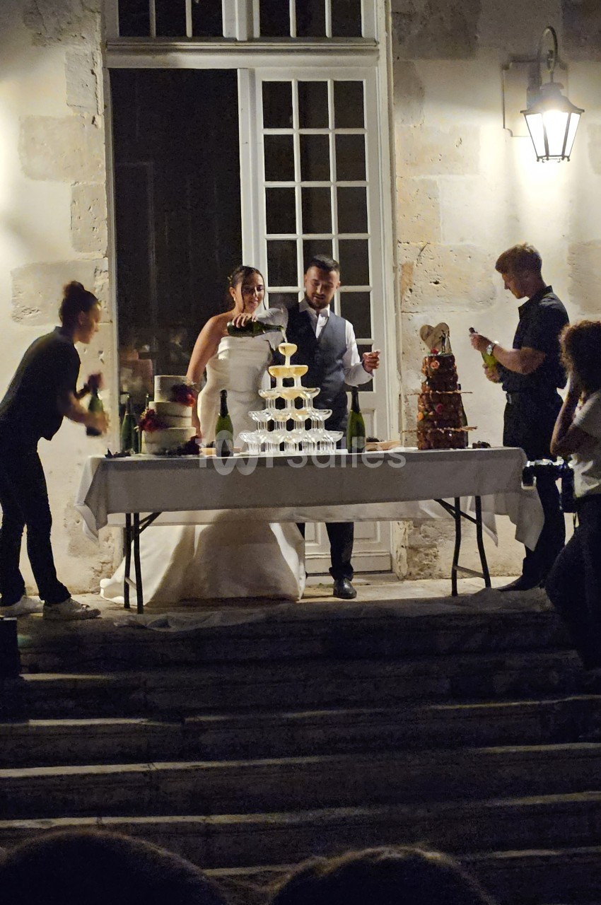 Un couple de mariés devant une pièce montée et des bouteilles de champagne, entouré de personnes en extérieur.