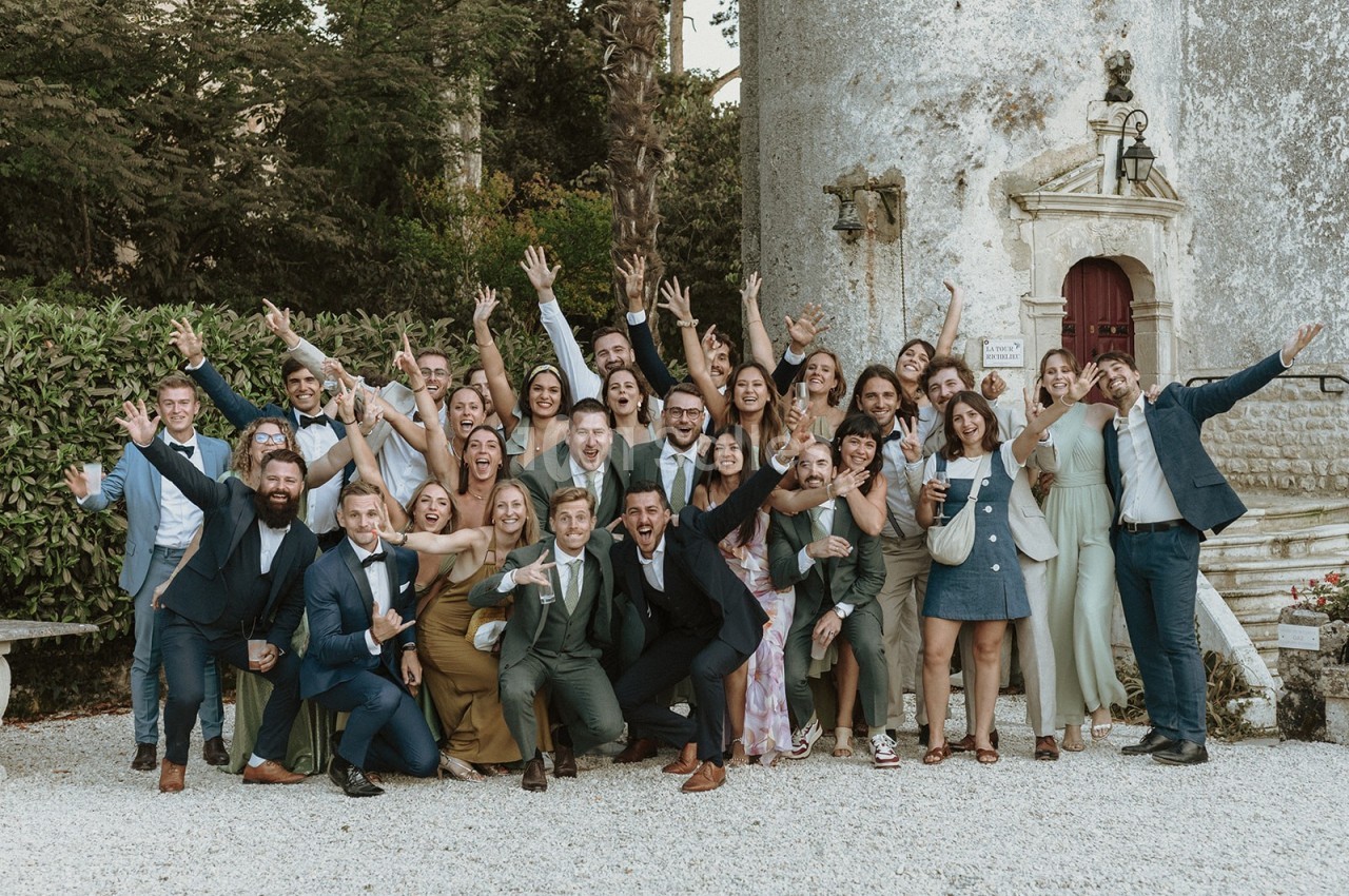 Un grand groupe de personnes souriantes posant devant un bâtiment ancien, bras levés dans une ambiance festive.
