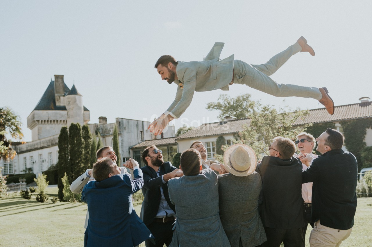 Un groupe d'hommes en costume lance un homme en l'air dans un jardin devant un bâtiment ancien.