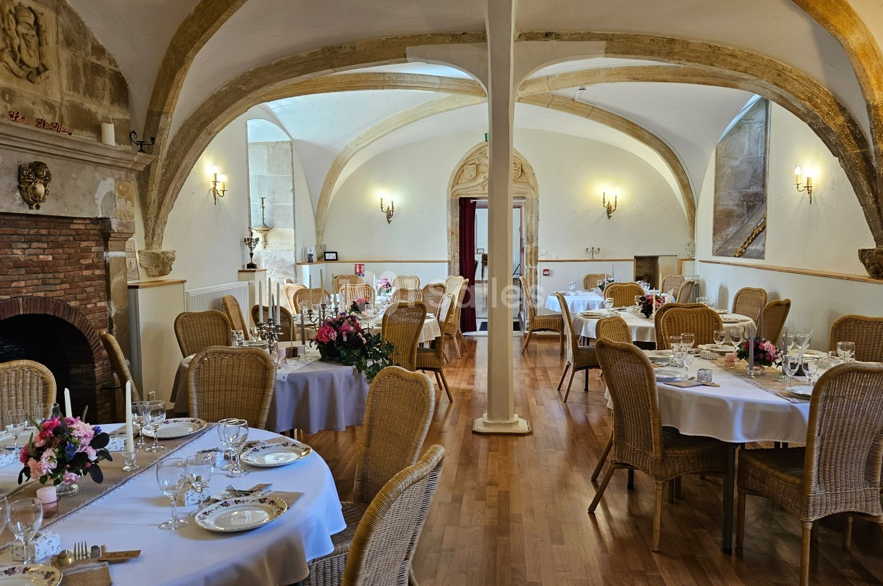 Salle de réception élégante avec tables dressées, chaises en osier, poutres apparentes et cheminée en pierre.