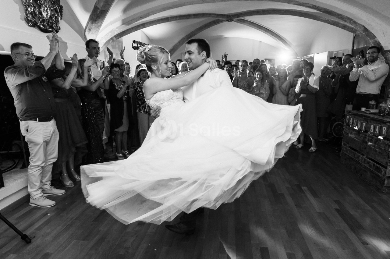 Un couple de mariés danse au centre d'une salle entourée d'invités applaudissant, sous des arches en pierre.