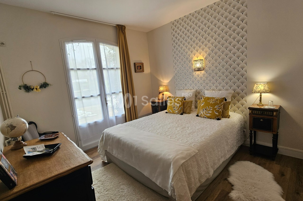 Chambre lumineuse avec lit double, coussins jaunes, mur capitonné, meubles en bois et fenêtre avec rideaux.