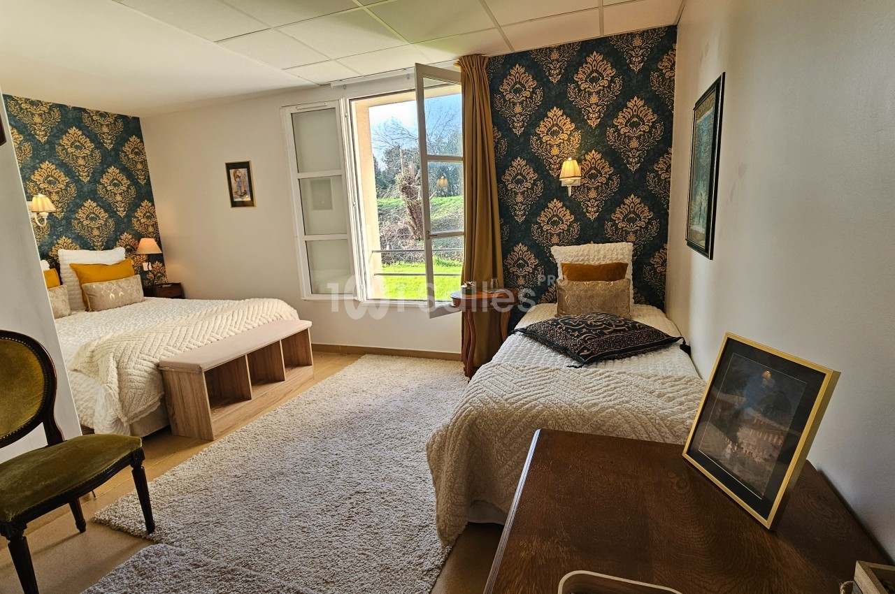 Chambre lumineuse avec deux lits simples, murs décorés de papier peint à motifs, fenêtre ouverte sur un paysage verdoyant.