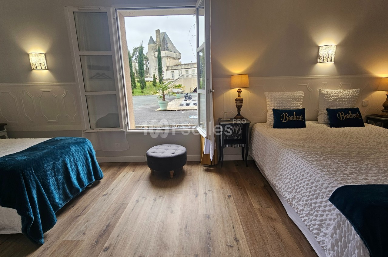 Chambre lumineuse avec deux lits, parquet, décoration élégante et vue sur un bâtiment historique par une fenêtre ouverte.