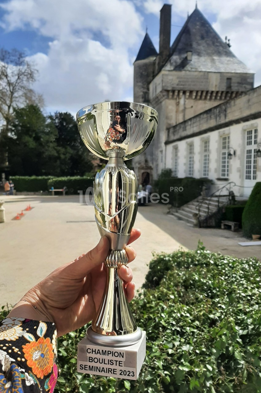 Une main tient un trophée devant un bâtiment historique entouré de verdure.