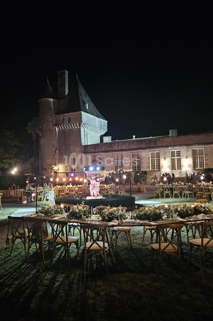 Vue nocturne d'un château éclairé, avec des tables décorées et des chaises disposées pour un événement en plein air.