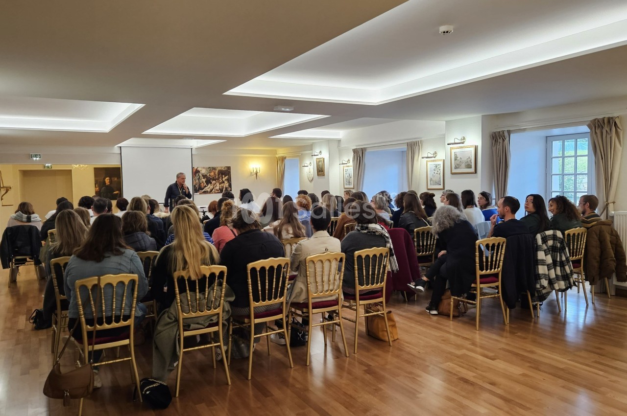 Un groupe de personnes assises dans une salle lumineuse, écoutant une présentation à l'avant.