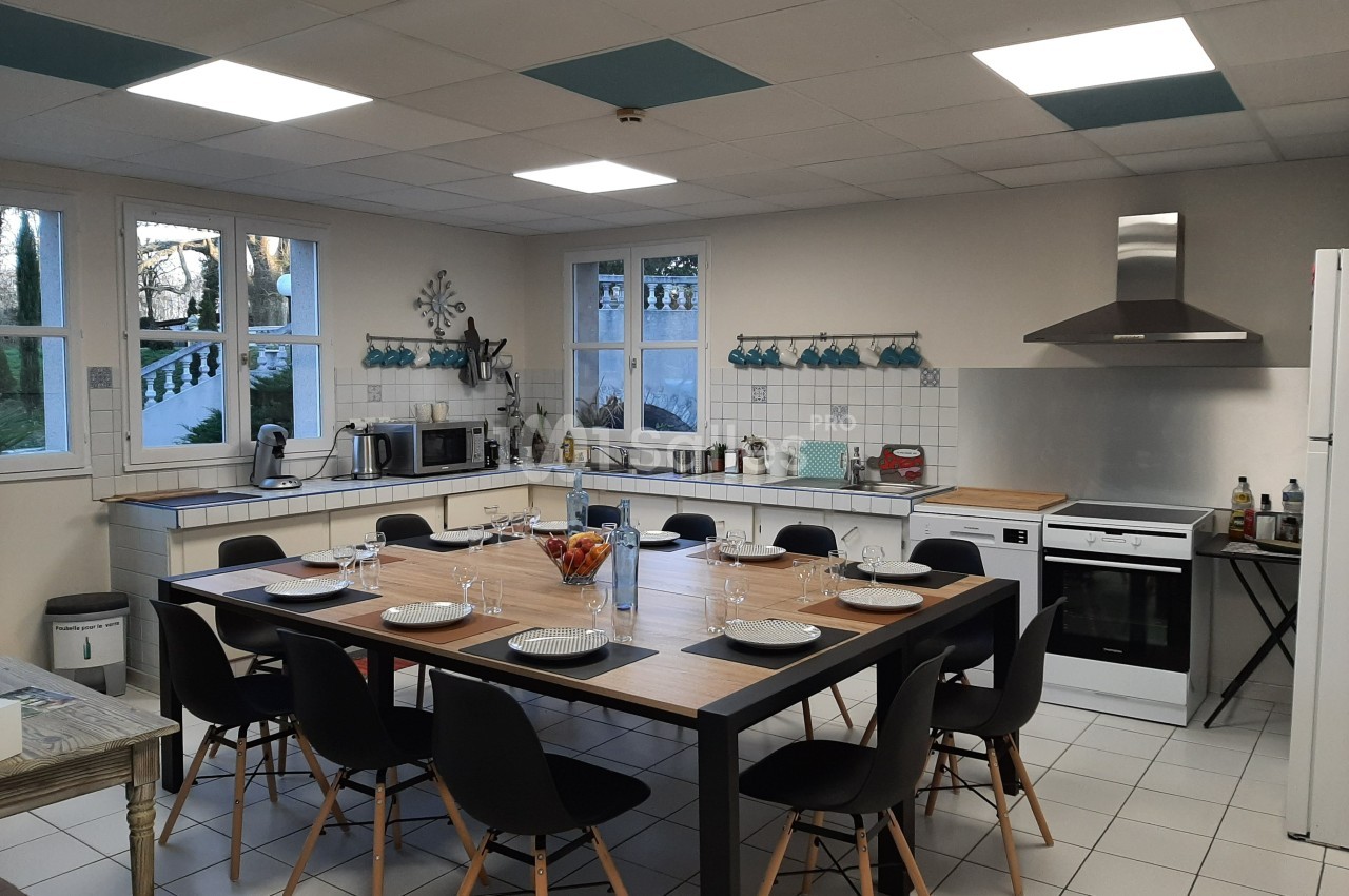 Cuisine spacieuse avec grande table dressée pour huit personnes, équipée d'appareils modernes et éclairée par des fenêtres.