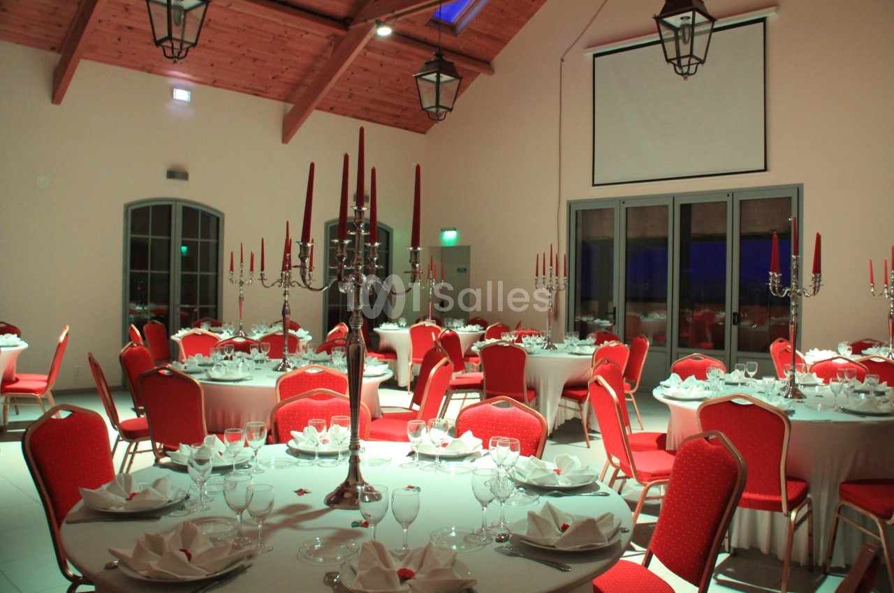 Salle de réception avec tables rondes dressées, chaises rouges et chandeliers argentés sous un plafond en bois.