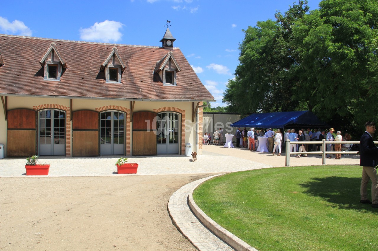 Bâtiment en briques avec toit en ardoise, terrasse pavée et groupe de personnes rassemblées sous un auvent bleu.