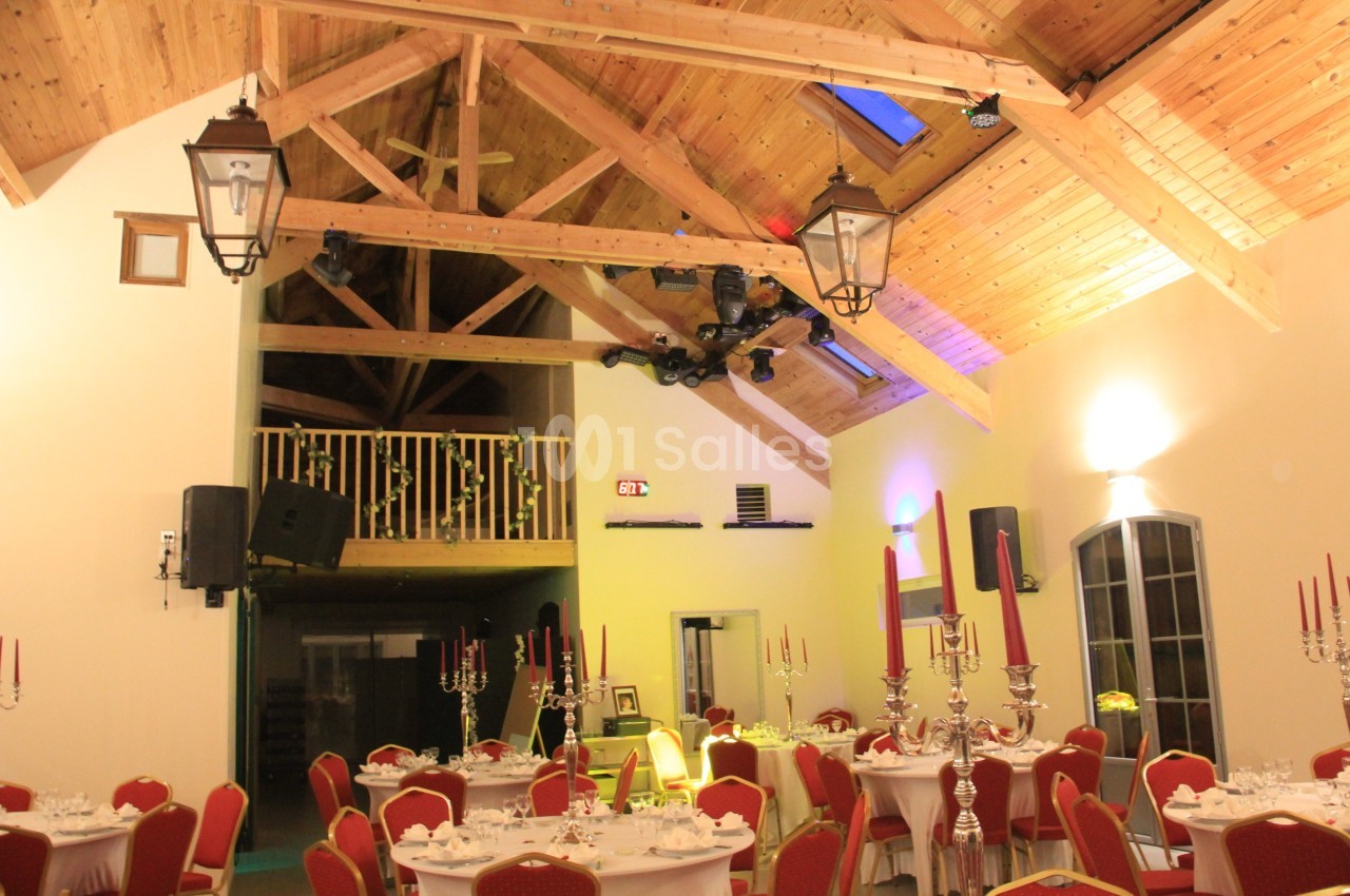 Salle de réception avec plafond en bois, tables dressées, chandeliers rouges et balcon intérieur.