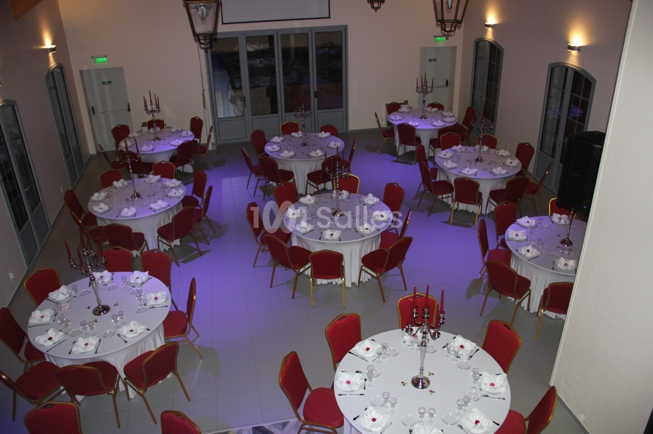 Salle de réception avec tables rondes dressées, nappes blanches, chaises rouges et chandeliers centraux.