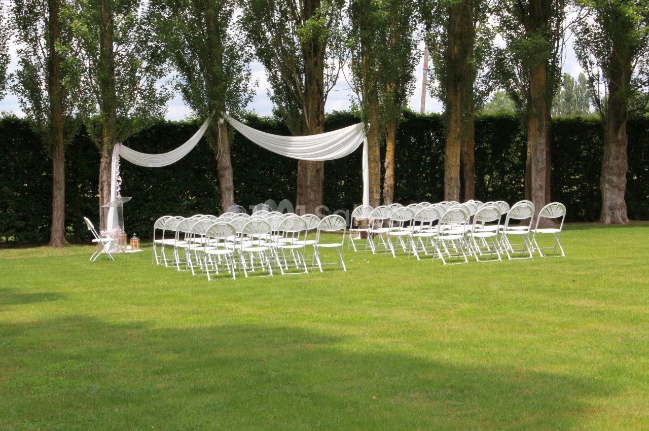 Chaises blanches disposées en rangées sur une pelouse, avec une arche décorée de tissu blanc devant des arbres.