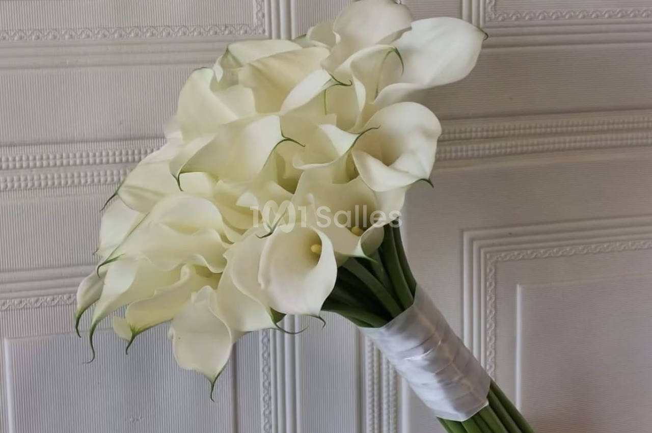 Bouquet de callas blancs élégamment disposés, attachés avec un ruban blanc, posé devant un mur texturé.