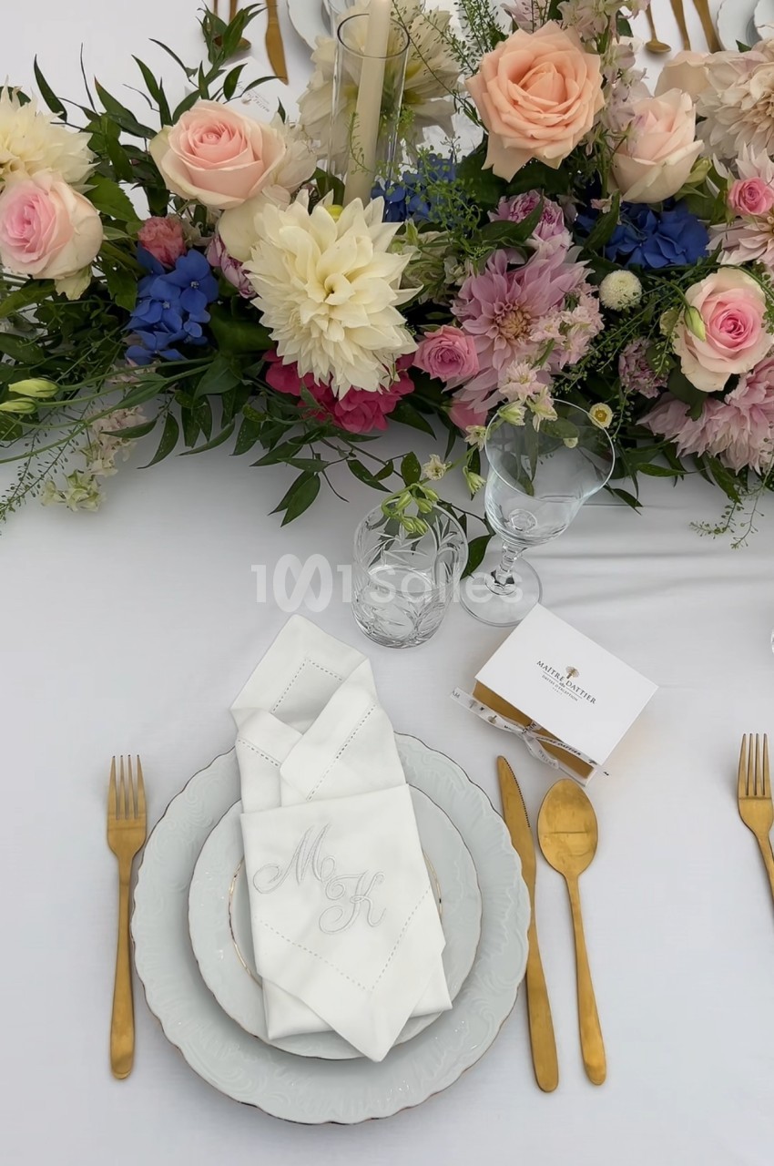 Table dressée avec vaisselle blanche, couverts dorés, serviette pliée et centre de table fleuri aux tons pastel.
