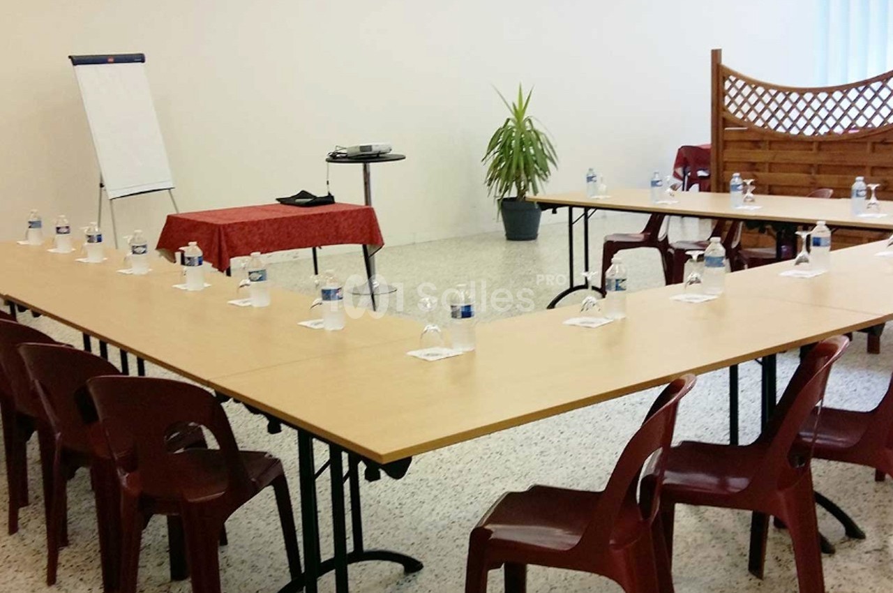 Salle de réunion avec tables disposées en U, chaises en plastique, tableau de conférence et plante en arrière-plan.