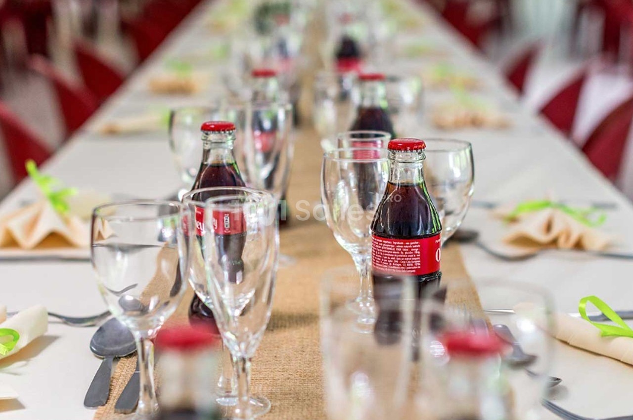Table dressée avec des verres, bouteilles de soda et serviettes pliées, décorée d’un chemin de table en toile de jute.