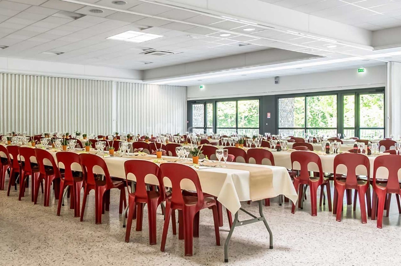Salle de réception lumineuse avec des tables dressées, nappes blanches et chaises rouges, prête pour un événement.