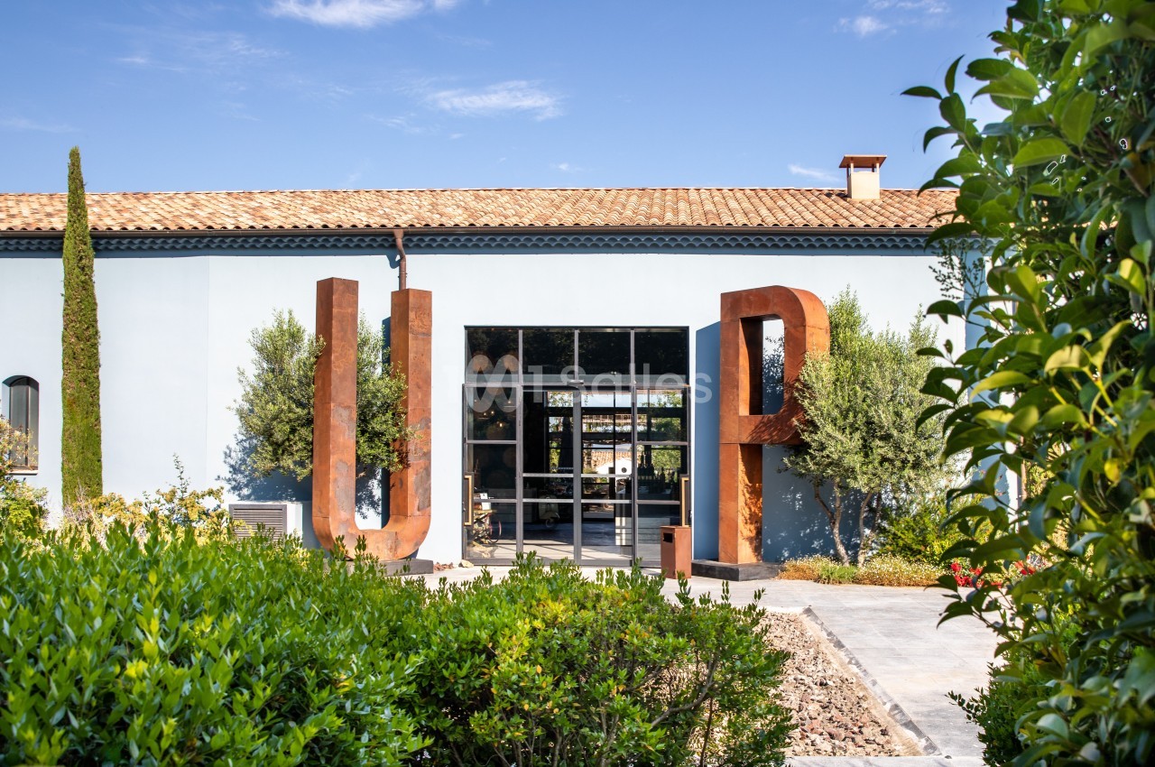 Façade d'un bâtiment moderne avec des lettres métalliques ’U’ et ’P’ devant une entrée vitrée, entourée de végétation.