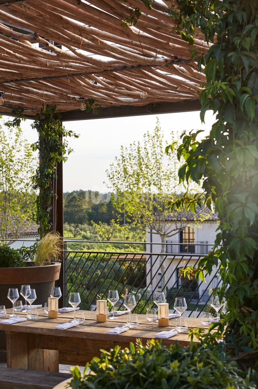 Table en bois dressée avec des verres et des lanternes sous une pergola en bois entourée de verdure, vue sur un paysage…