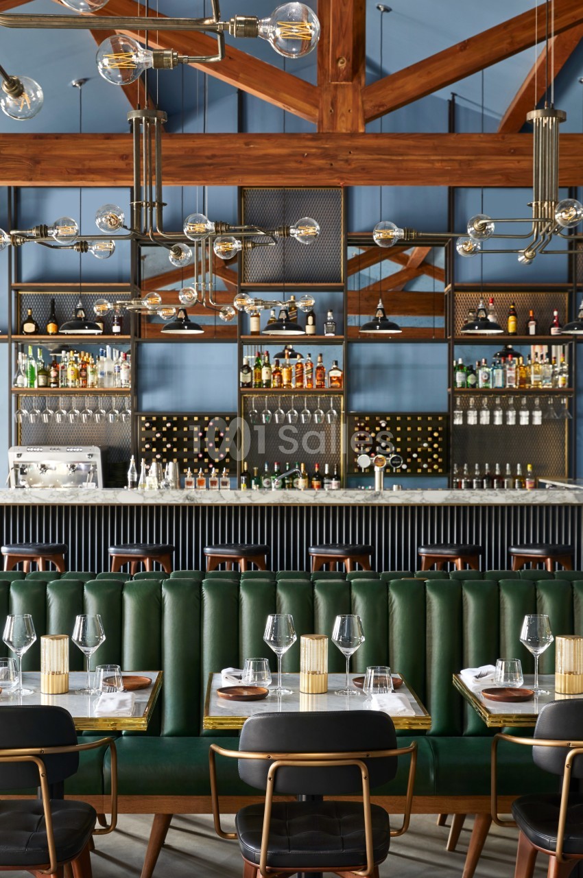 Bar élégant avec étagères remplies de bouteilles, banquettes vertes, tables dressées et luminaires suspendus modernes.