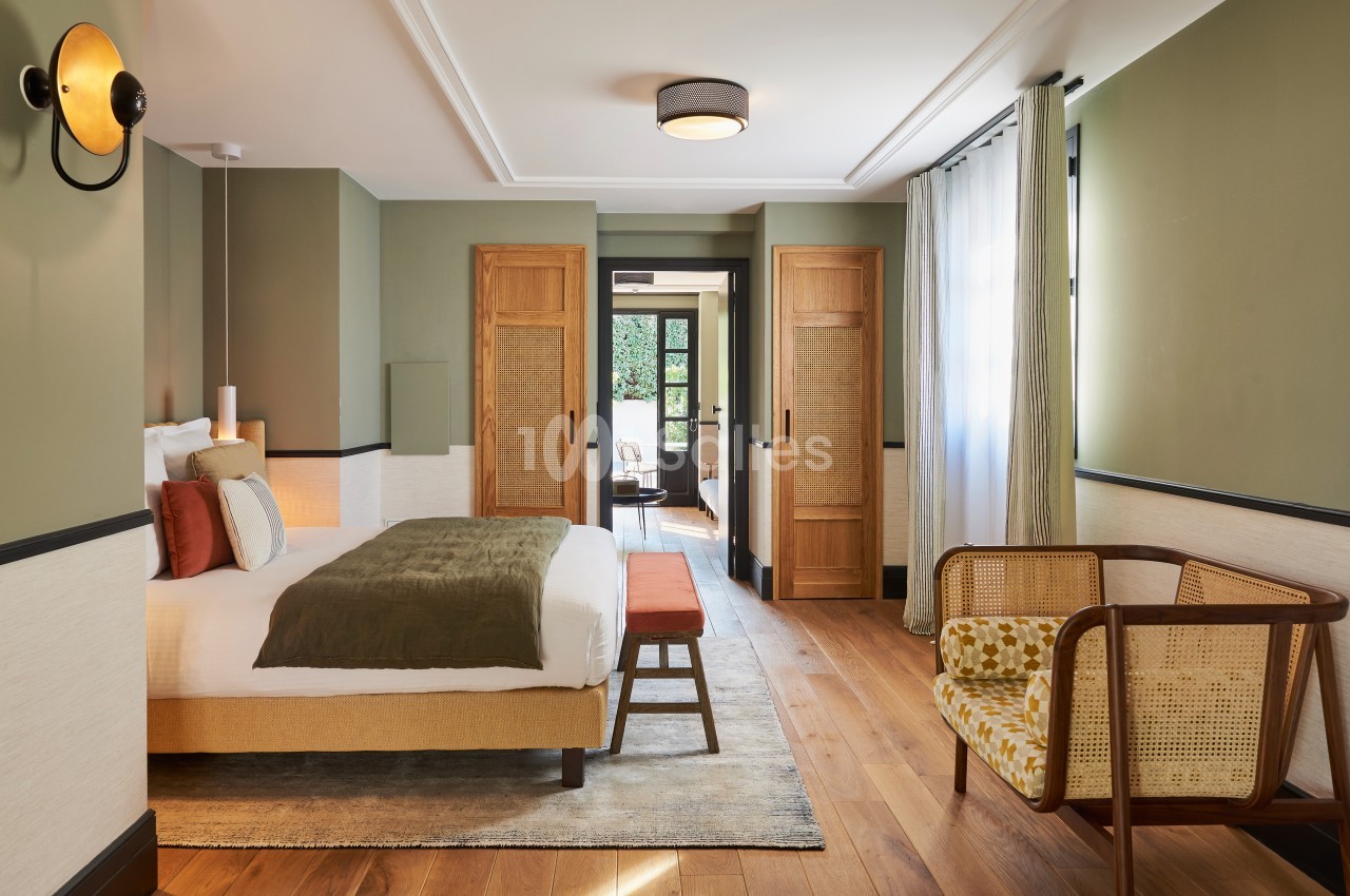 Chambre lumineuse avec lit double, fauteuil en rotin, parquet en bois et portes en bois donnant sur une terrasse.