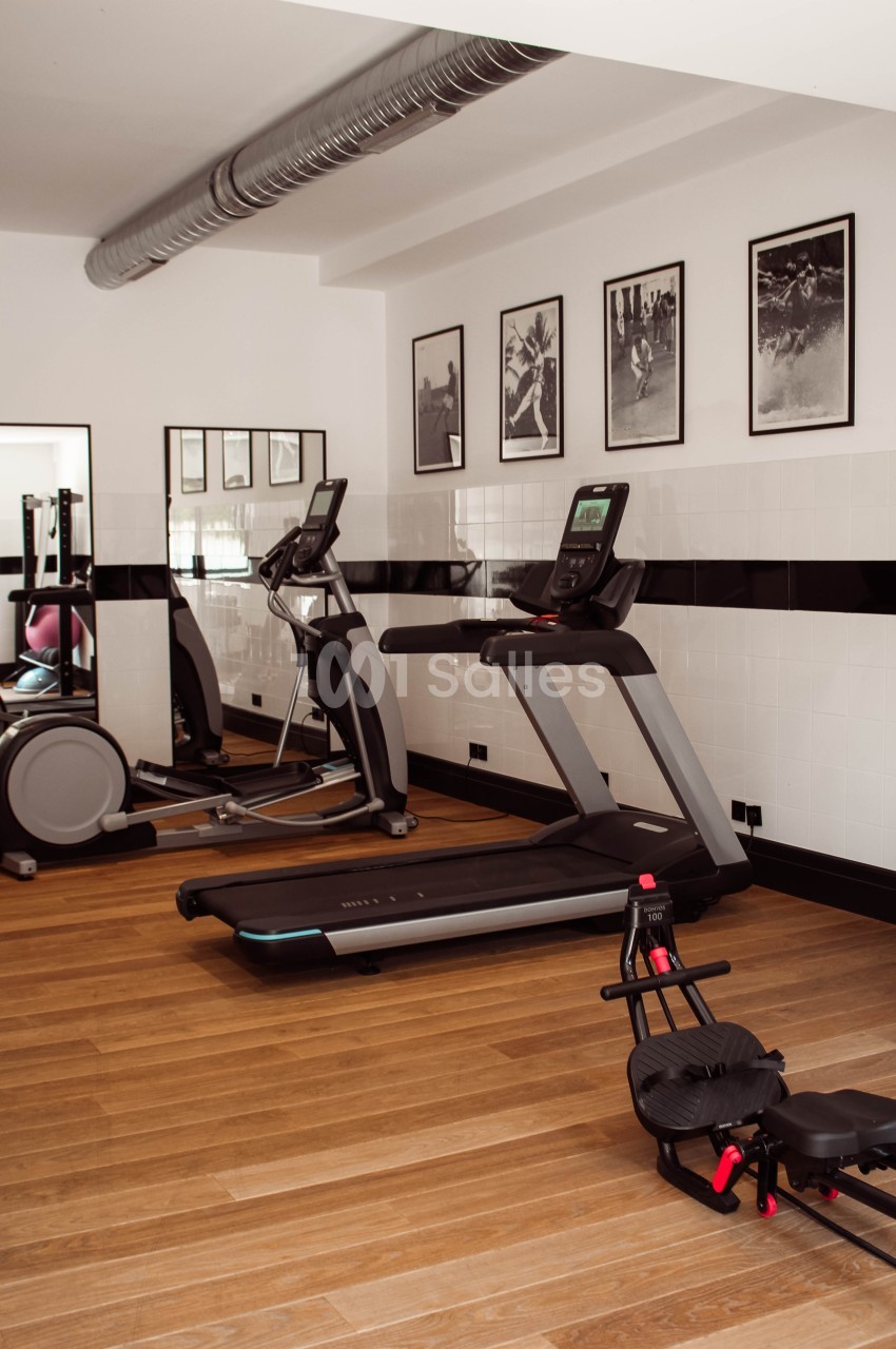 Salle de sport avec tapis de course, vélo elliptique, rameur et décorations murales en noir et blanc.