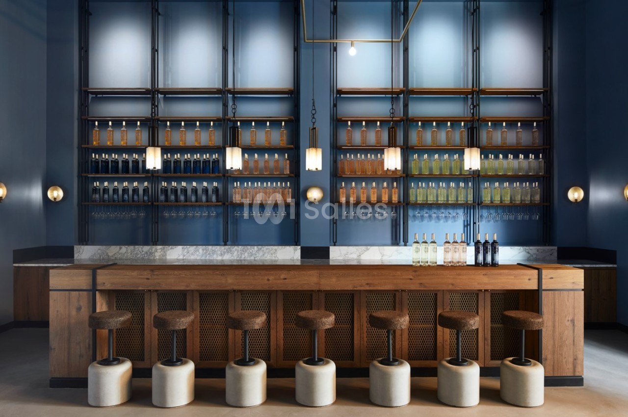 Bar moderne avec comptoir en bois, tabourets rembourrés et étagères murales garnies de bouteilles alignées.