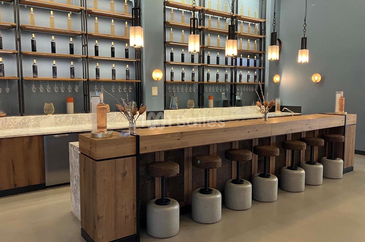 Bar moderne avec comptoir en bois, tabourets rembourrés, étagères murales garnies de bouteilles et éclairage suspendu.