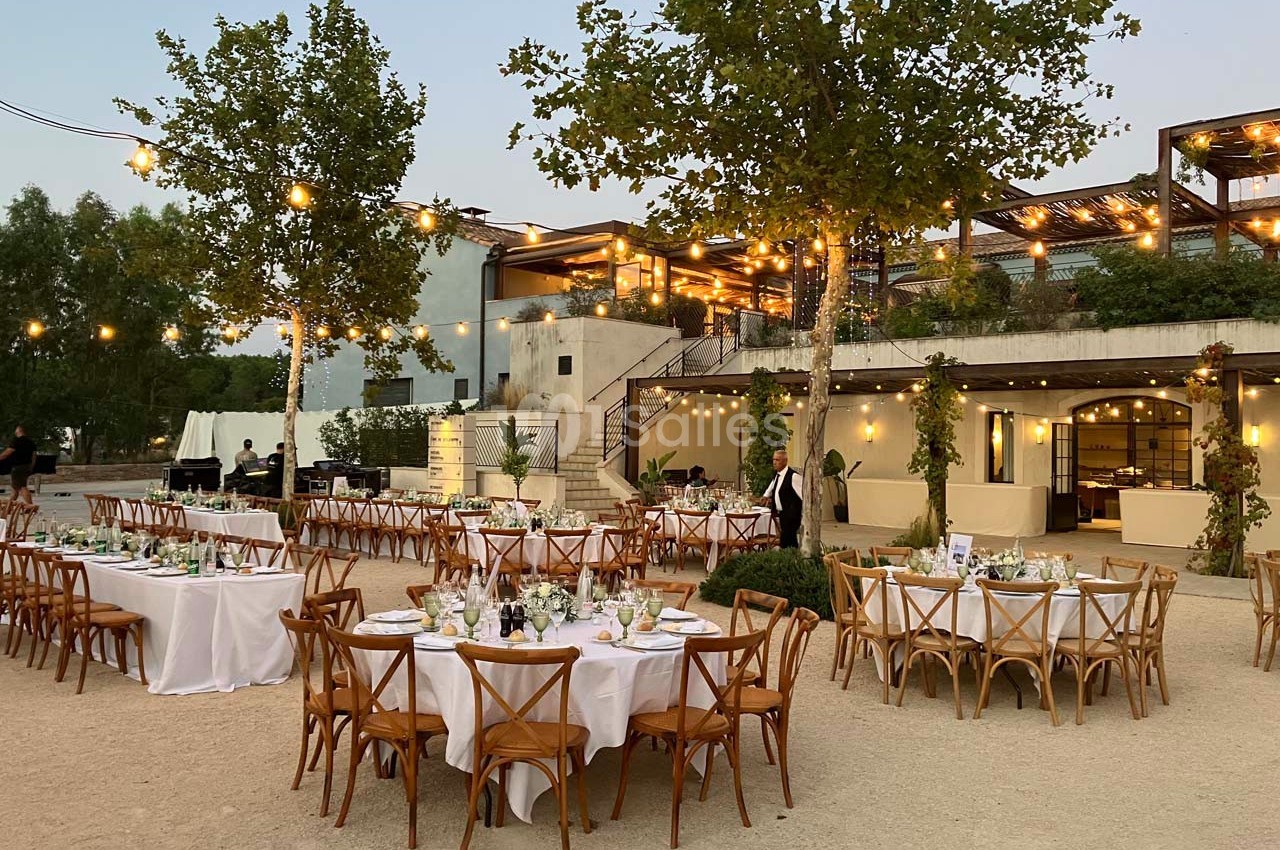 Tables rondes dressées avec nappes blanches dans un espace extérieur éclairé par des guirlandes lumineuses au crépuscule.