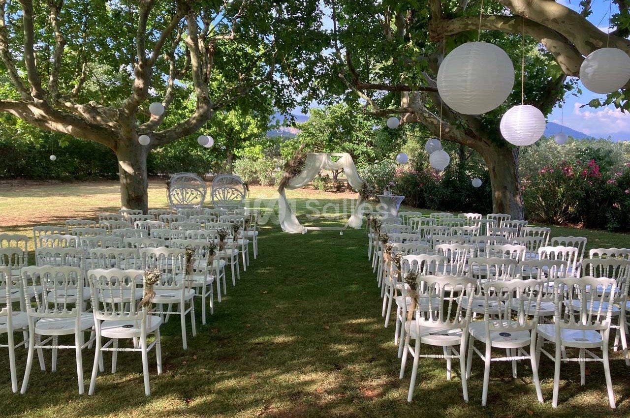 Chaises blanches alignées en extérieur sous des arbres, avec une arche décorée et des lanternes suspendues.
