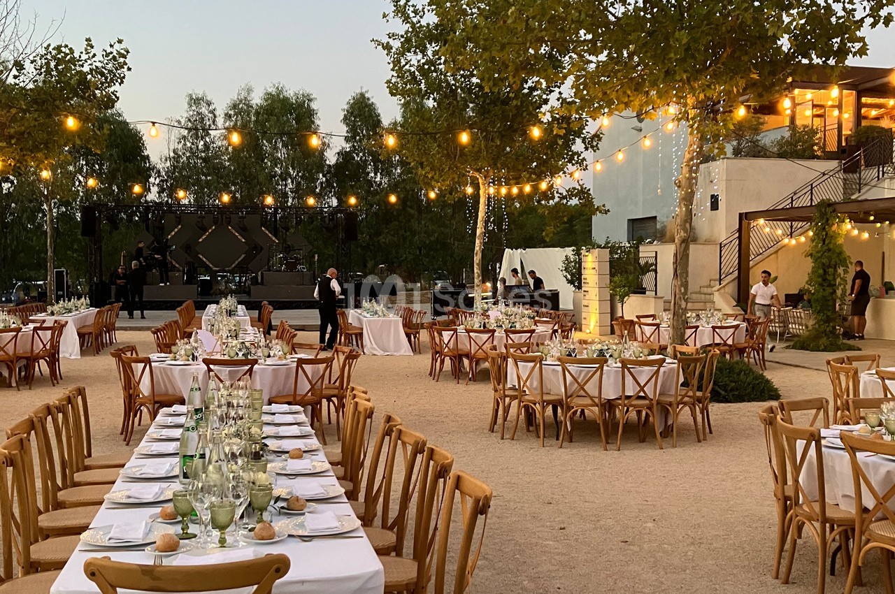 Tables dressées en extérieur pour un événement, entourées de guirlandes lumineuses et de végétation au crépuscule.