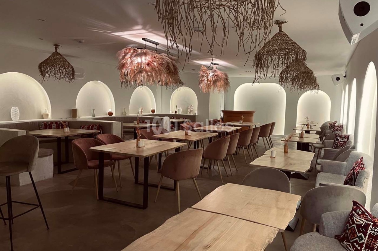 Salle de restaurant lumineuse avec tables en bois clair, chaises rembourrées et suspensions en fibres naturelles.