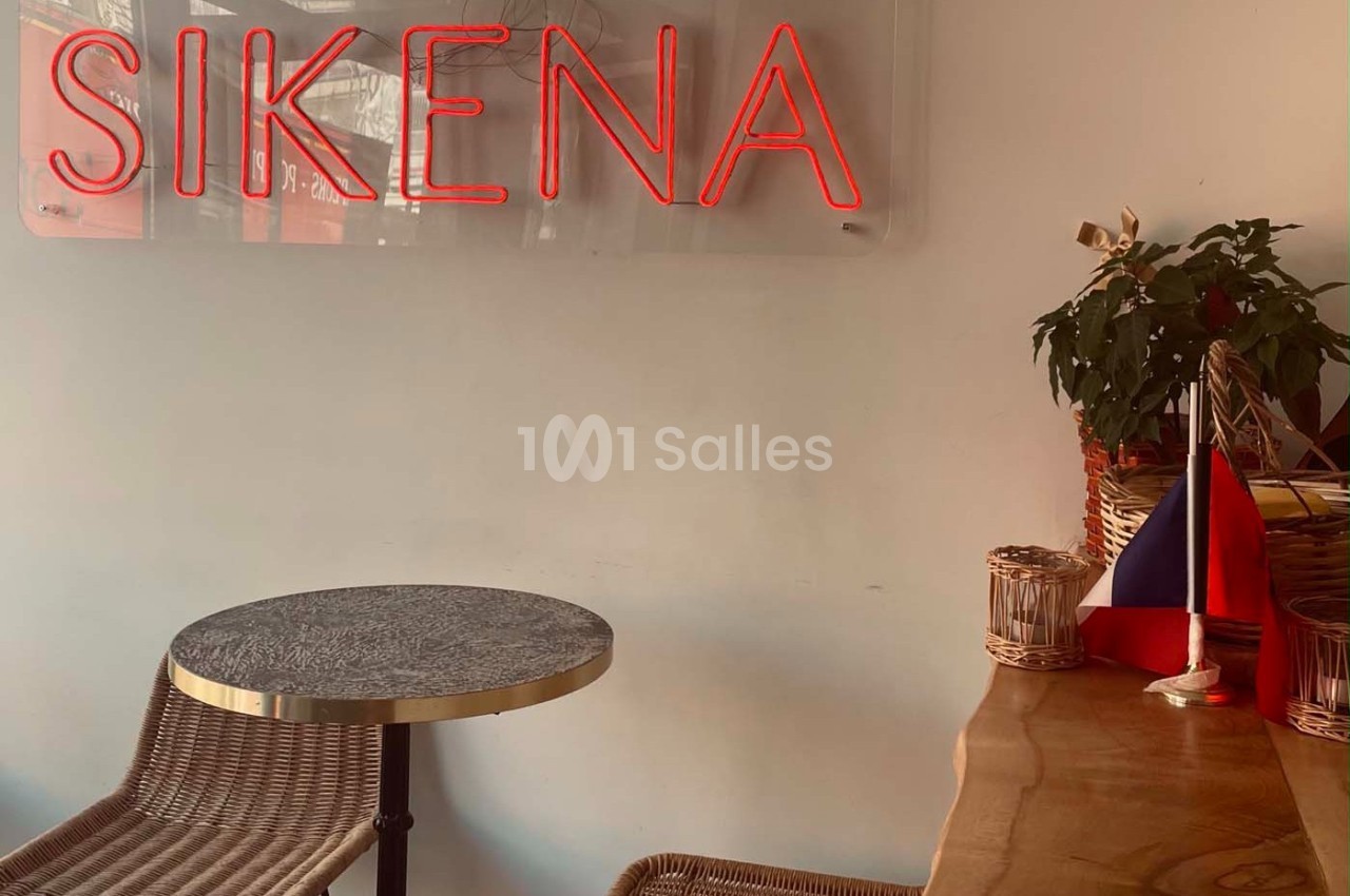 Table et chaises en rotin dans un café avec une enseigne lumineuse rouge ’SIKENA’ et un drapeau sur le comptoir.
