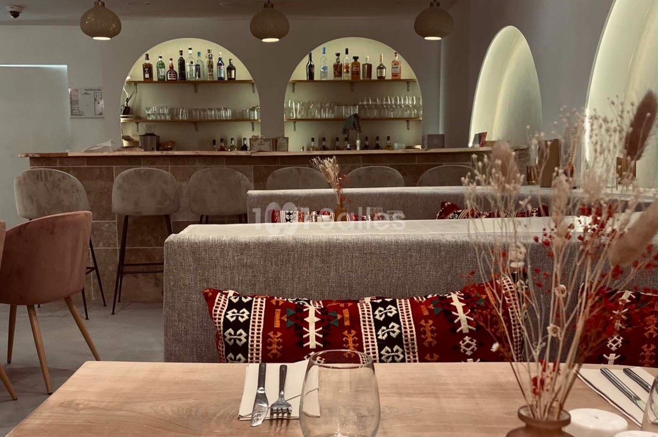 Salle de restaurant avec tables dressées, banquettes ornées de coussins à motifs, et un bar en arrière-plan.
