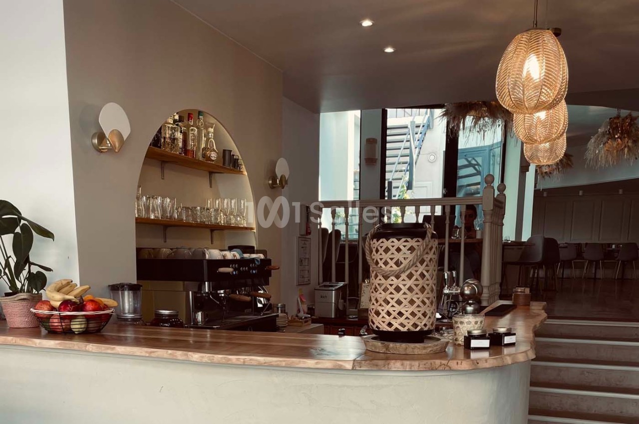 Comptoir de café avec machine à expresso, étagères de verres et bouteilles, éclairage chaleureux et décoration en rotin.