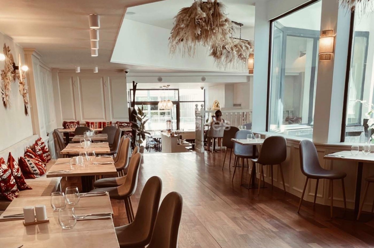 Salle de restaurant lumineuse avec tables en bois, chaises modernes, grandes fenêtres et décorations suspendues.