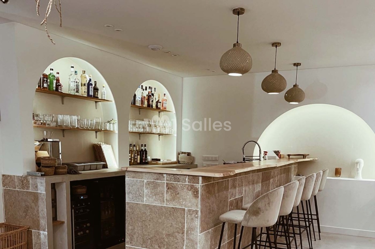 Bar moderne avec comptoir en bois, tabourets rembourrés, étagères éclairées contenant des bouteilles et des verres.
